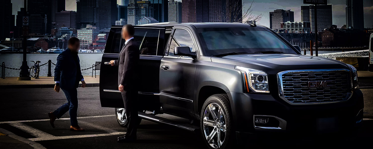 Security opening door of 2018 GMC Yukon Denali for client in parking lot in front of Boston skyline